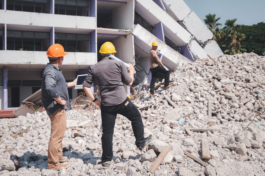 Three construction workers wearing safety helmets stand on a pile of rubble, evaluating a partially collapsed building during a demolition project.